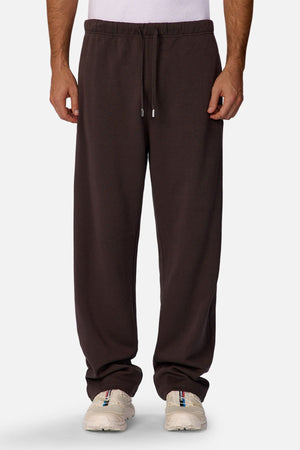 Wearing the Industrie The Astoria Lux Tech Trackpant in loose dark brown for premium comfort, paired with white sneakers and a white shirt, a person stands against a plain light background, showcasing effortless casual style.