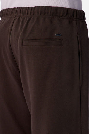 Close-up of the back of a person wearing Industrie The Astoria Lux Tech Trackpant in dark brown, featuring relaxed fit, elastic waistband, back pocket, and a small rectangular metal logo above the pocket for premium comfort.