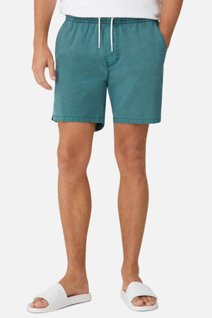 Wearing Industrie The Bahama Angler Shorts in teal, featuring an elastic waist and white drawstring, hands casually in pockets. The outfit is completed with a crisp white shirt and matching slide sandals against a simple backdrop, highlighting the relaxed style.