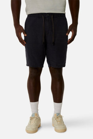 A person wearing a light gray shirt, Industrie The Baller Linen Short in dark blue with a drawstring waist and relaxed fit, and white socks with cream-colored sneakers featuring light blue accents stands against a plain background.