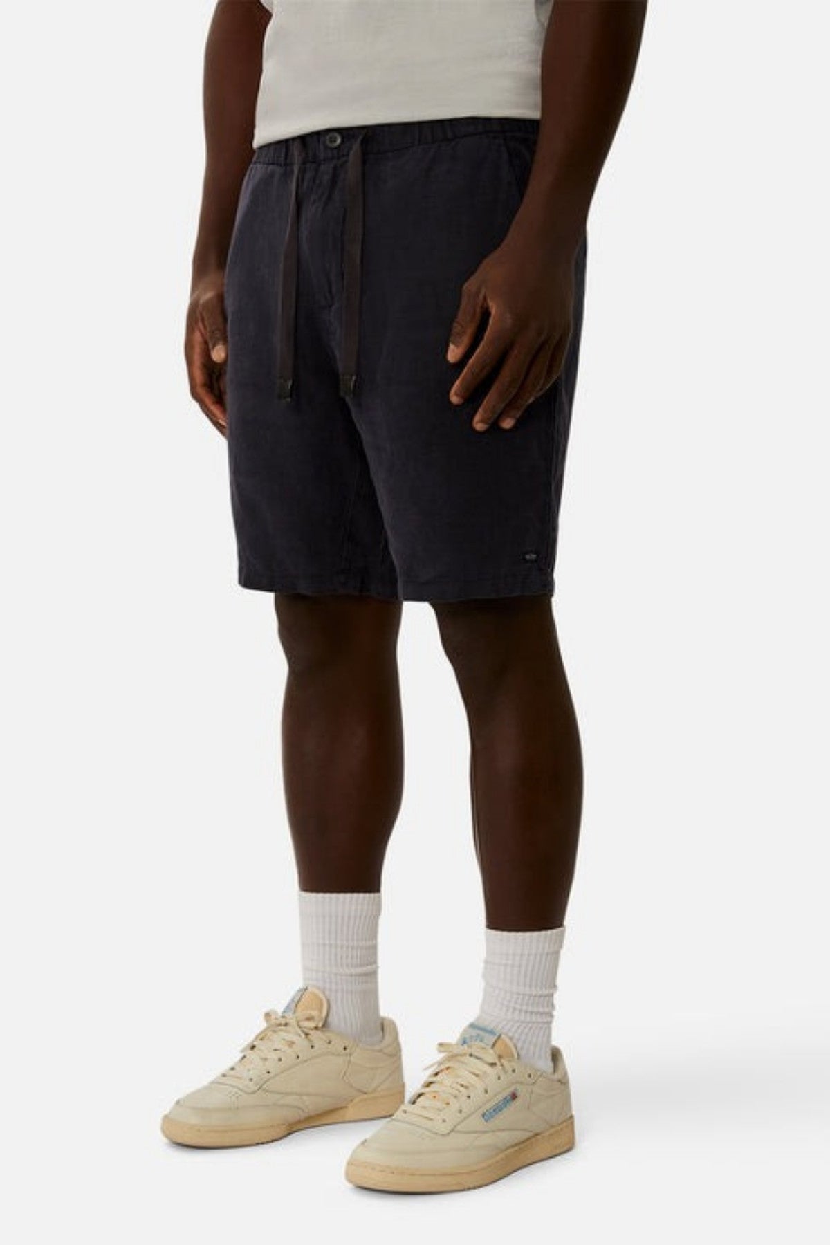 A person is wearing Industrie The Baller Linen Short in dark navy, featuring a drawstring waist that's visible. They have paired it with white socks and beige sneakers, standing against a plain light background, showing only the lower half of their body.