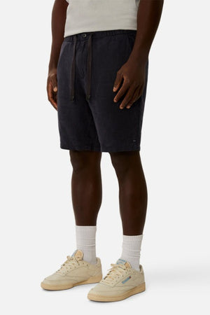 A person is wearing Industrie The Baller Linen Short in dark navy, featuring a drawstring waist that's visible. They have paired it with white socks and beige sneakers, standing against a plain light background, showing only the lower half of their body.