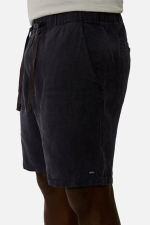 Side view of a person wearing the Industrie The Baller Linen Short, which are knee-length and feature a dark color with an elastic drawstring waist. The fabric has a smooth texture, and there is a small logo at the bottom of the shorts. The background is plain white.