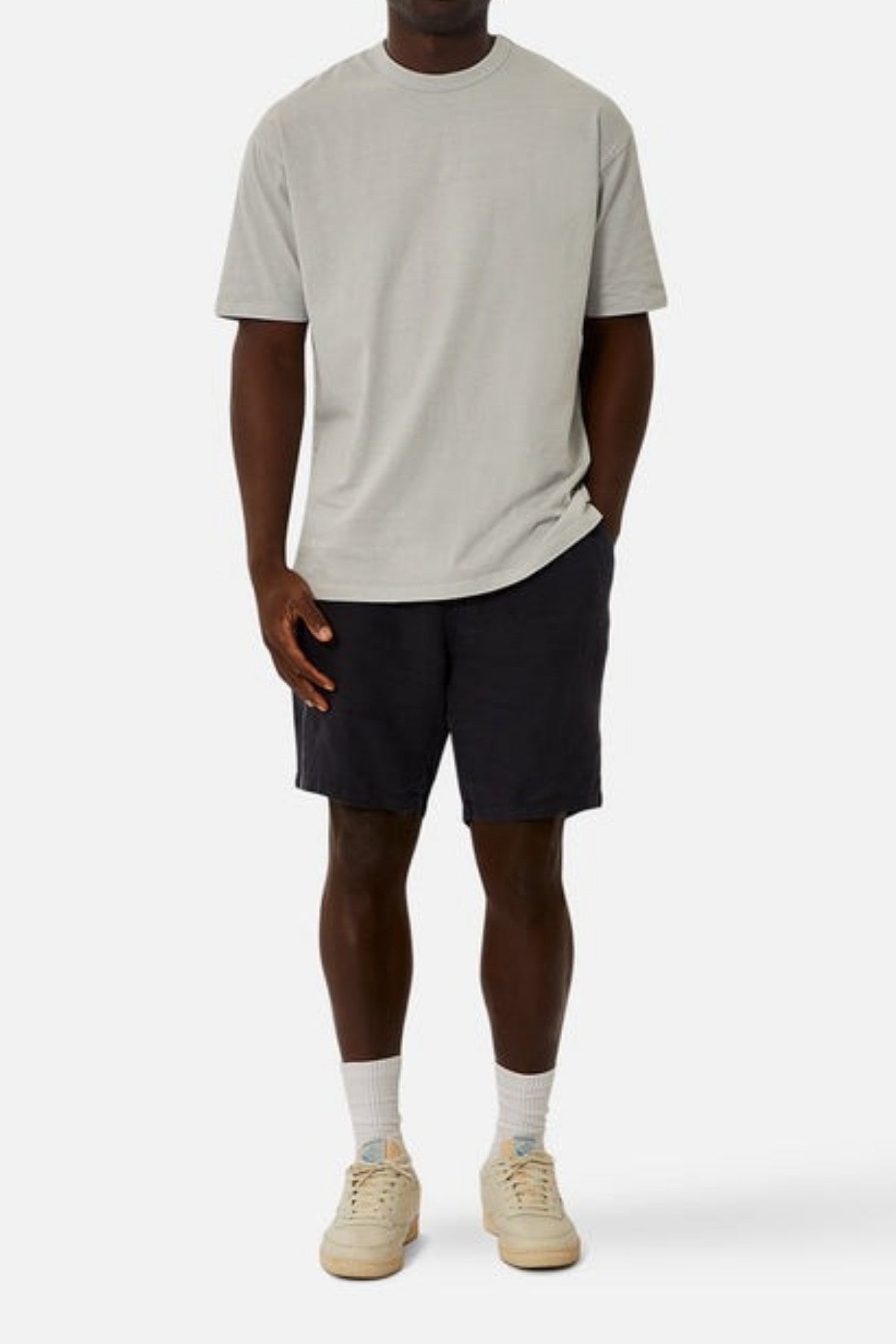 A person wearing a light gray T-shirt and the Industrie The Baller Linen Shorts, characterized by their knee-length cut and drawstring waist, paired with beige sneakers, stands against a plain white background. Their left hand is in their pocket as they look forward.