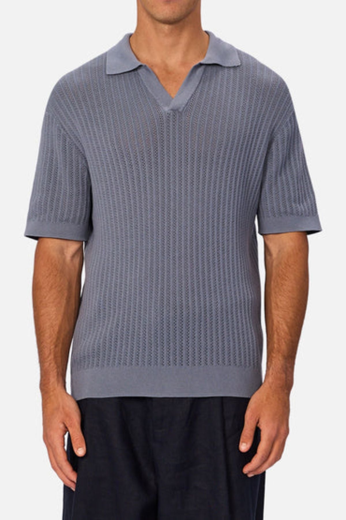 A man wearing Industrie The Bellano Polo—a short-sleeve, textured gray 100% cotton men's polo with a V-neck collar—and dark pants stands against a plain light background.