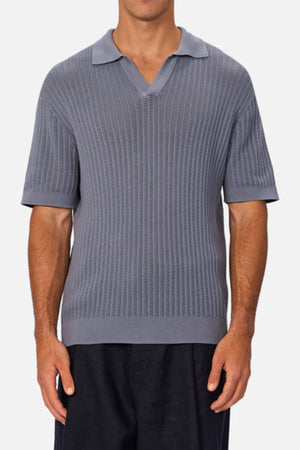 A man wearing Industrie The Bellano Polo—a short-sleeve, textured gray 100% cotton men's polo with a V-neck collar—and dark pants stands against a plain light background.
