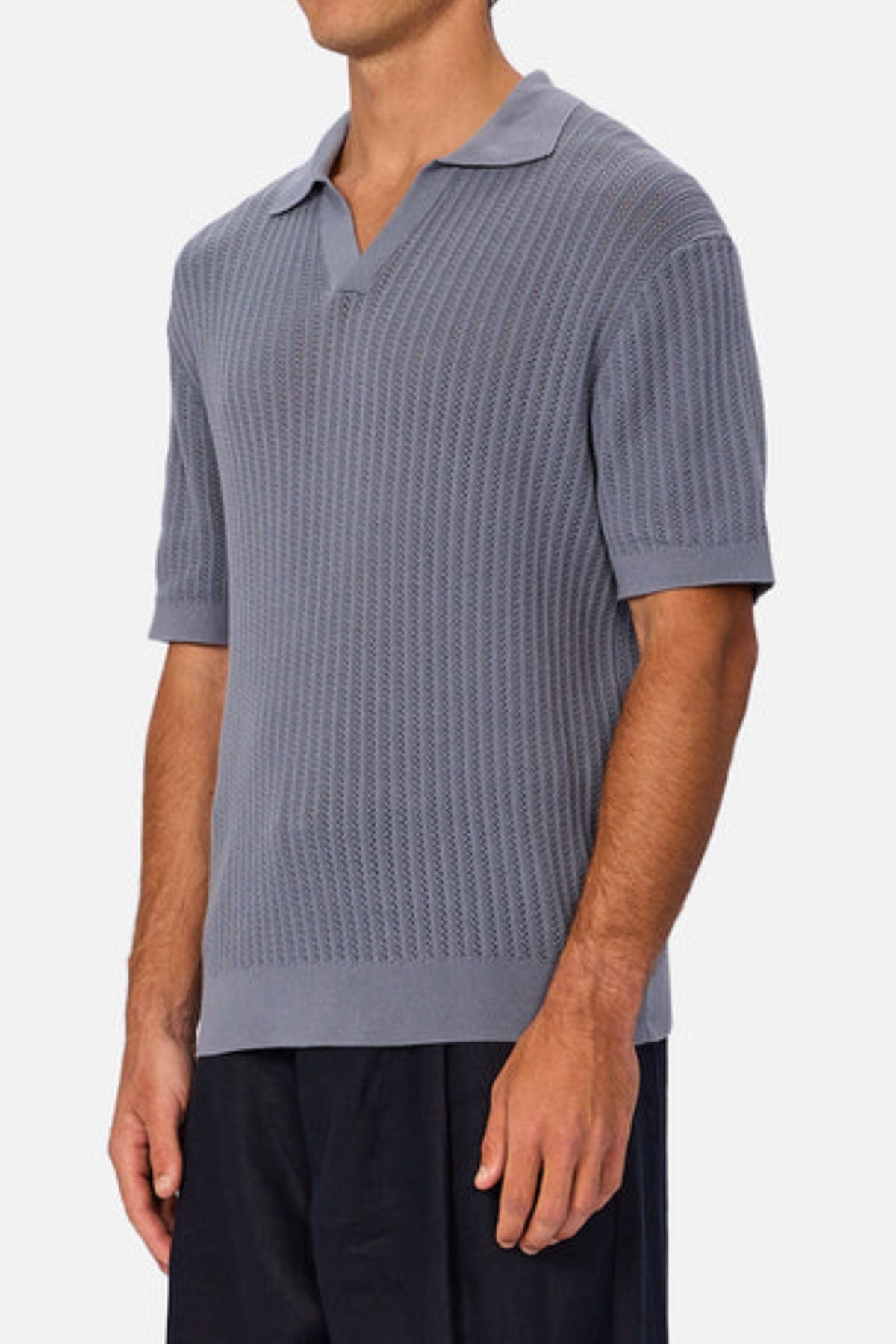 A man wearing the Industrie The Bellano Polo, a light blue 100% cotton knit polo with short sleeves, collar, and open neckline, paired with black pants, stands against a plain light background.