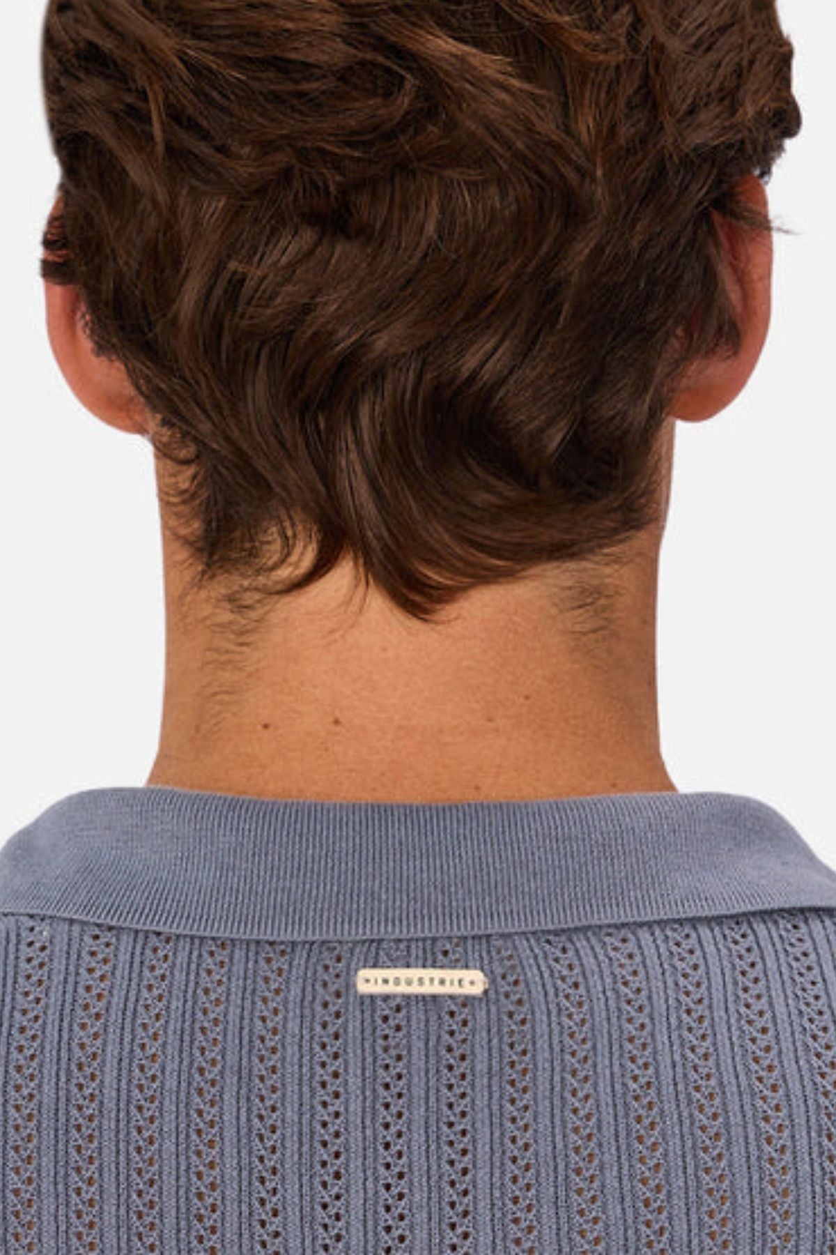 A person with short, wavy brown hair is seen from behind, wearing the Industrie The Bellano Polo in light blue, crafted from 100% cotton with a textured knit and a small "INDUSTRIE" label on the back collar.