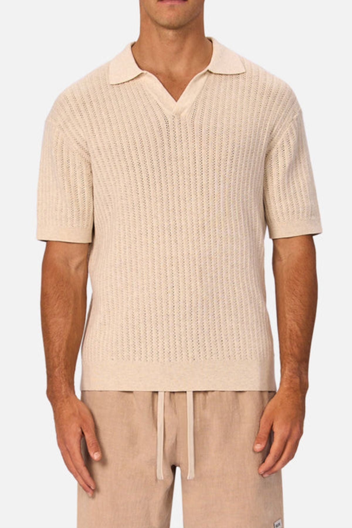 A man wearing the Industrie The Bellano Polo, a light beige short-sleeve knit with a textured pattern and collar, paired with light tan drawstring shorts, stands against a plain white background.