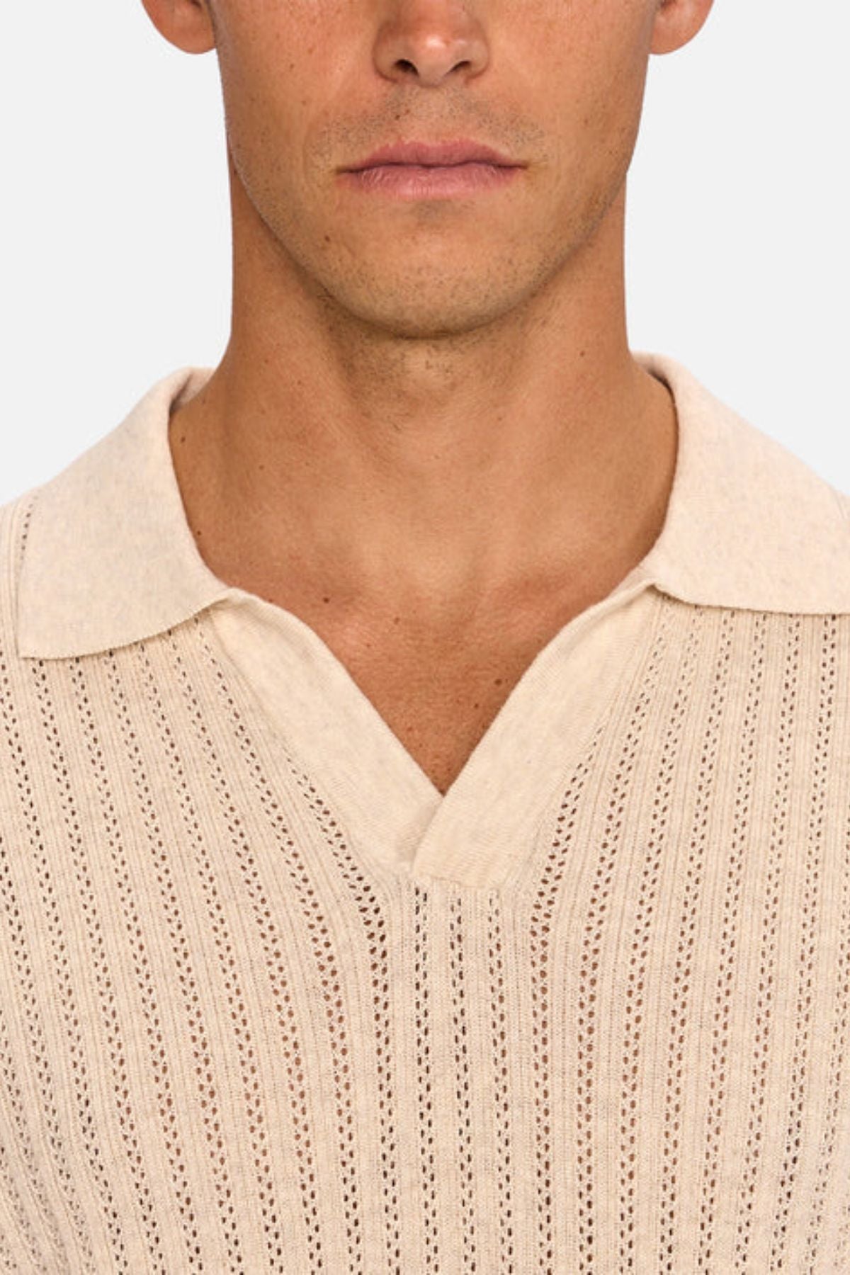 A close-up of a person wearing the Industrie The Bellano Polo, a beige men's short-sleeve knit polo with collar and V-neckline. Made from breathable cotton with a vertical perforated pattern. Only the lower half of the face is shown.