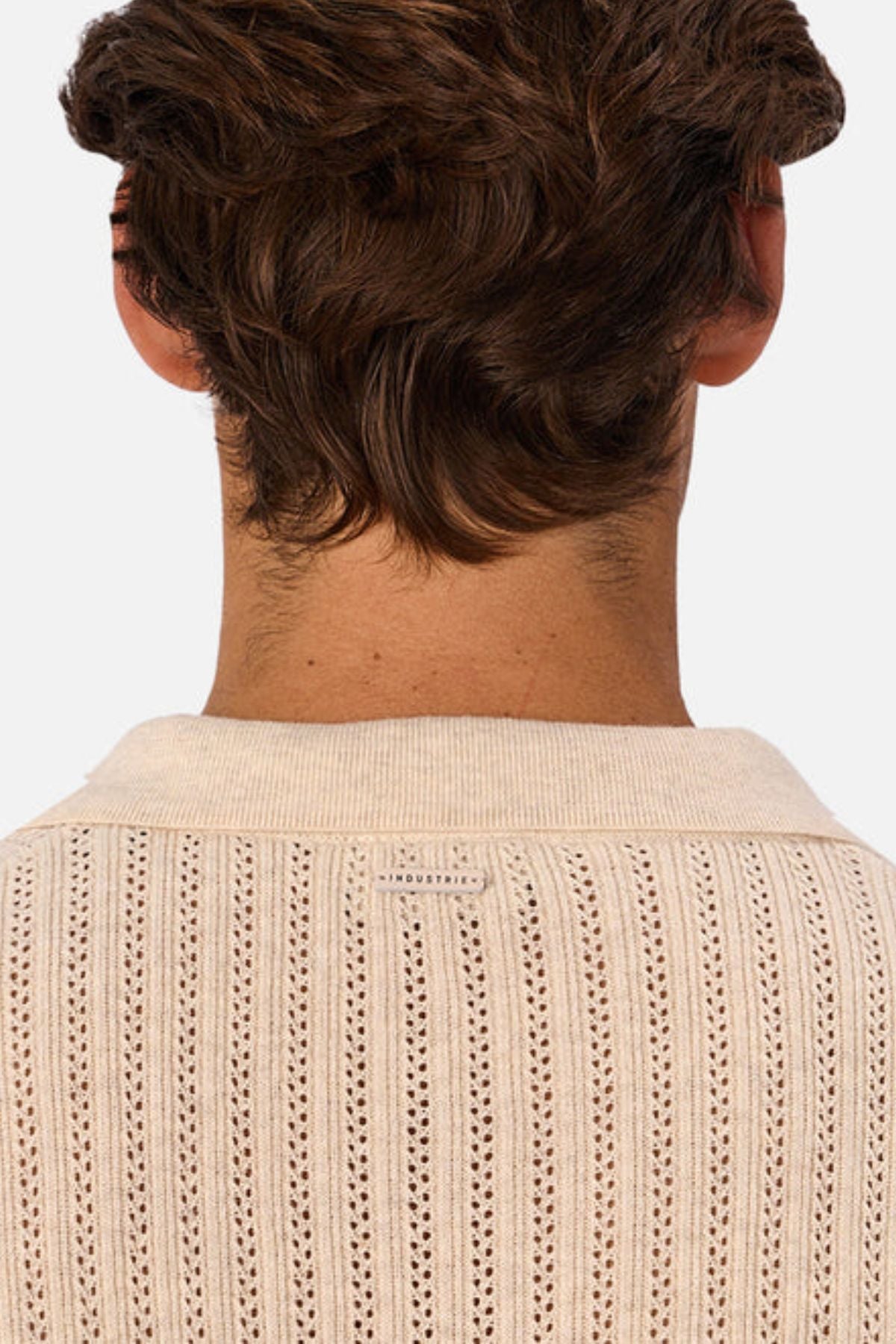 The image shows the back of a person with short, wavy brown hair wearing a cream-colored Industrie The Bellano Polo men's knit sweater, featuring a small label at the top center of the back.