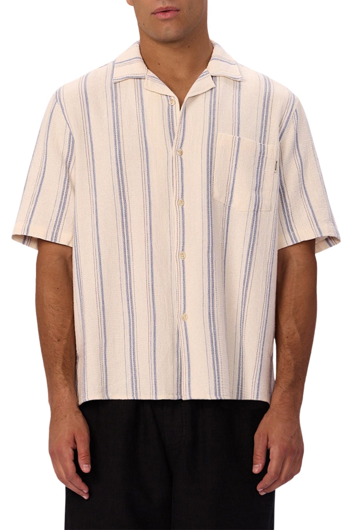 A man wears the Industrie The Bowment Short Sleeve Shirt—cream with blue vertical stripes, textured fabric, front pocket—paired with black pants, making it a stylish choice for any casual wardrobe.