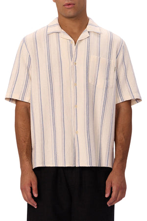 A man wears the Industrie The Bowment Short Sleeve Shirt—cream with blue vertical stripes, textured fabric, front pocket—paired with black pants, making it a stylish choice for any casual wardrobe.
