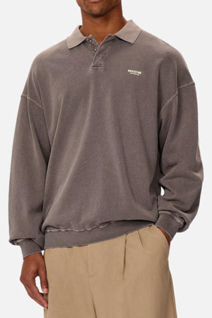 Someone wearing a loose-fit, faded gray Industrie The Burbank Polo Track shirt with a collar and small white chest text, paired with beige pleated pants. The image is cropped at the neck and knees.
