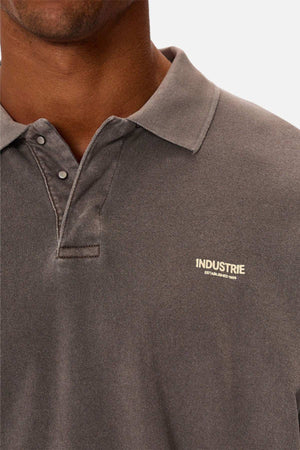 A close-up shows someone wearing Industrie The Burbank Polo Track, a faded grey collared shirt with premium fabric, two buttons undone, and "INDUSTRIE" printed on the left chest; their face is partly visible.