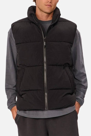 Someone stands with hands in pockets, wearing the Industrie The Busan Puffer Vestโa black nylon vest with Poly-fil paddingโover a gray long-sleeve shirt and dark pants, against a plain light background.