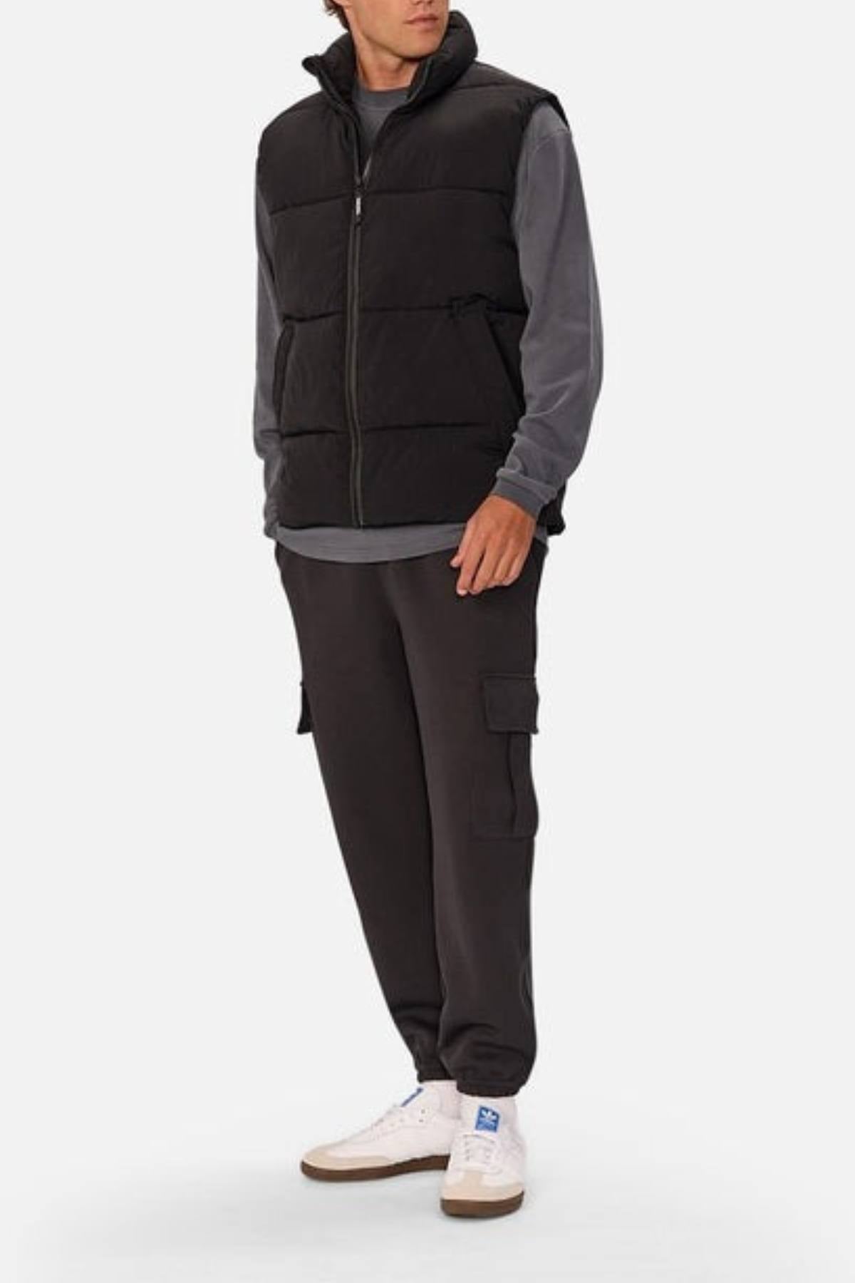 A person wears the Industrie The Busan Puffer Vest, a black nylon vest with Poly-fil padding, over a gray long-sleeve shirt, paired with black cargo pants and white sneakers, standing against a plain light background.