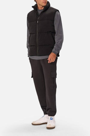 A person wears the Industrie The Busan Puffer Vest, a black nylon vest with Poly-fil padding, over a gray long-sleeve shirt, paired with black cargo pants and white sneakers, standing against a plain light background.