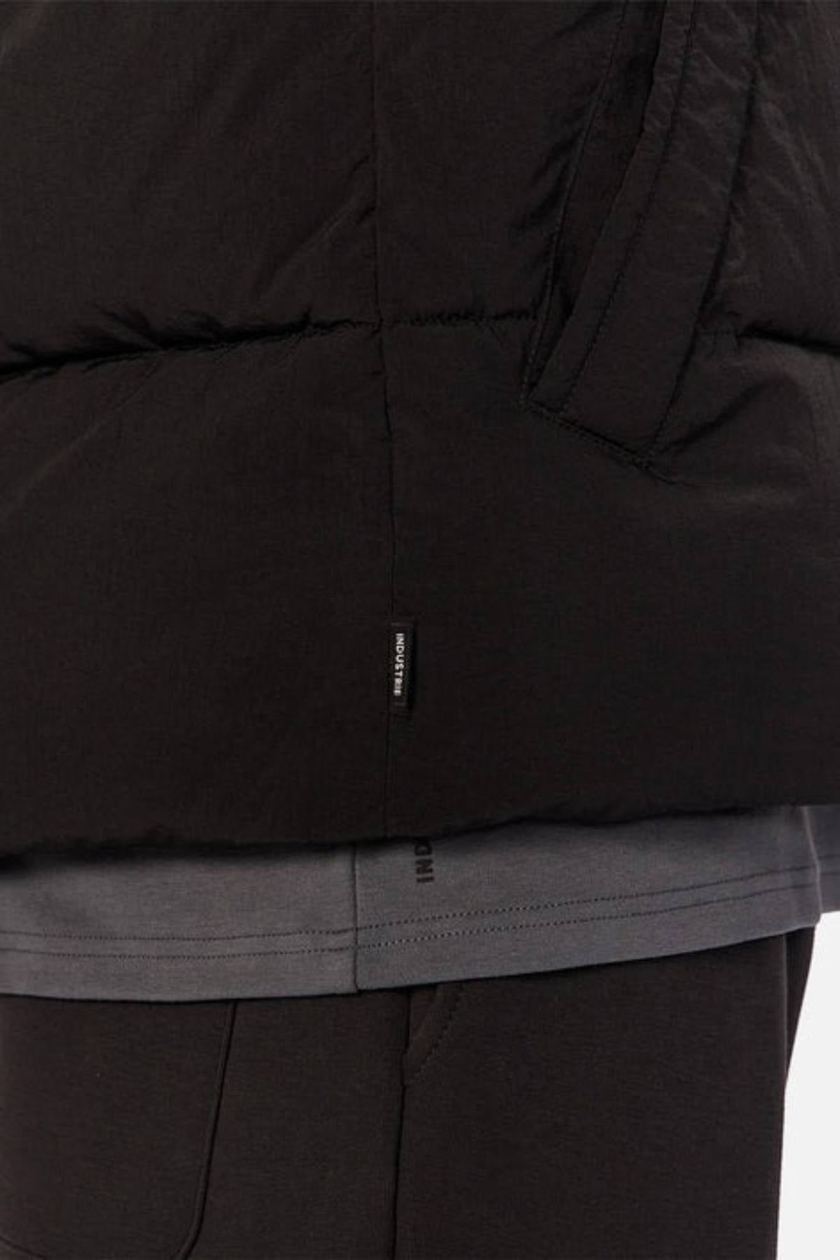 Close-up of a person wearing the Industrie The Busan Puffer Vest in black nylon with Poly-fil padding, visible pocket, and a small black tag with white text near the seam, paired with a grey t-shirt and black pants.