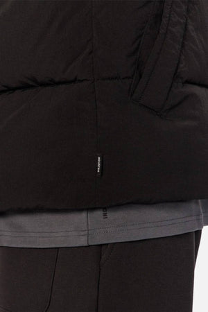 Close-up of a person wearing the Industrie The Busan Puffer Vest in black nylon with Poly-fil padding, visible pocket, and a small black tag with white text near the seam, paired with a grey t-shirt and black pants.
