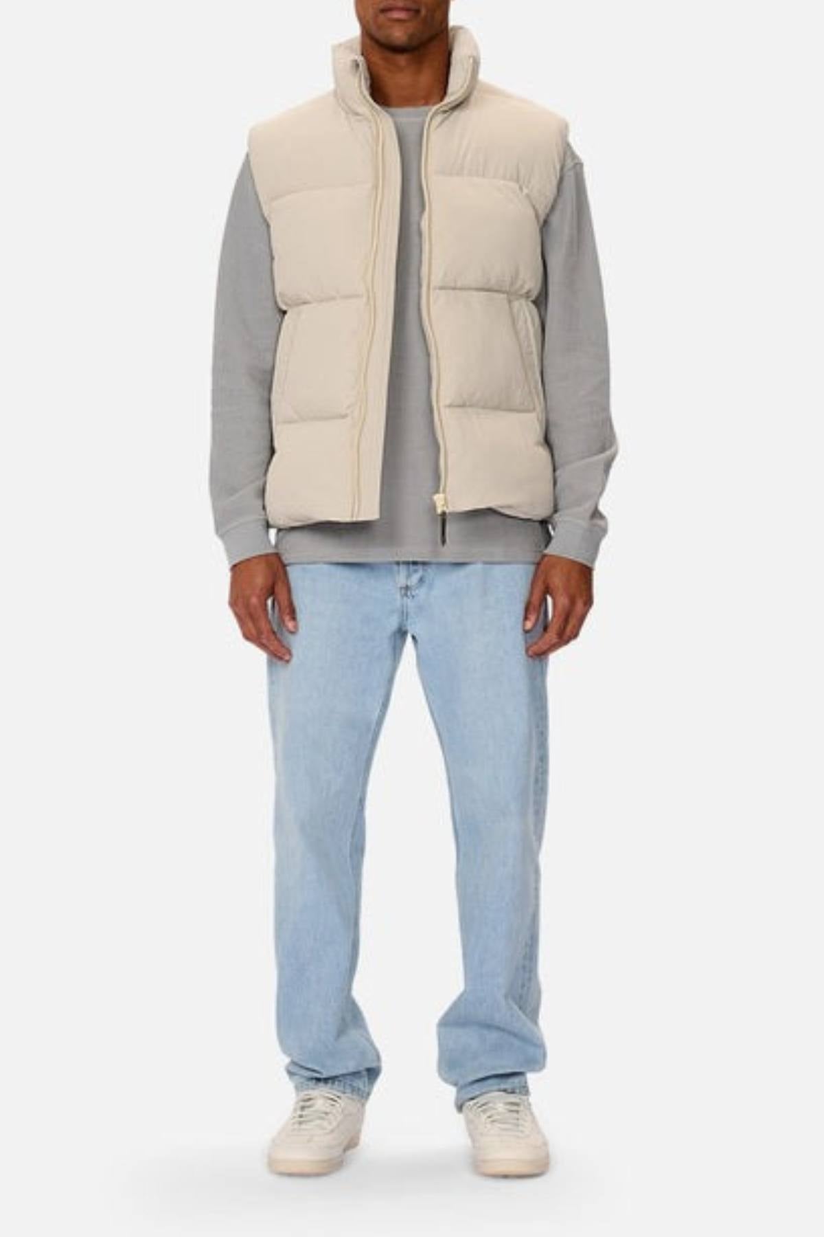 A person faces forward wearing the Industrie The Busan Puffer Vest in beige with Poly-fil Padding over a gray long-sleeve shirt, light blue jeans, and white sneakers. Their head is not fully visible against a plain light background.