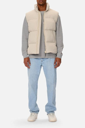 A person faces forward wearing the Industrie The Busan Puffer Vest in beige with Poly-fil Padding over a gray long-sleeve shirt, light blue jeans, and white sneakers. Their head is not fully visible against a plain light background.