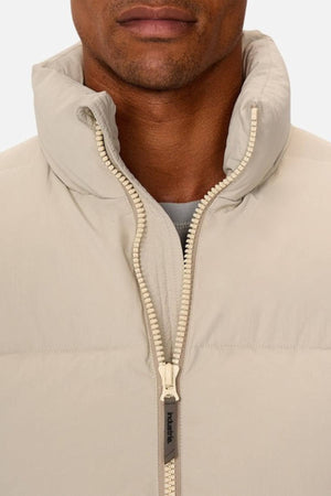 A close-up shows a person wearing the Industrie The Busan Puffer Vest in light beige nylon with Poly-fil padding, a high collar, and partially unzipped front revealing a gray shirt. Only the lower half of their face is visible.