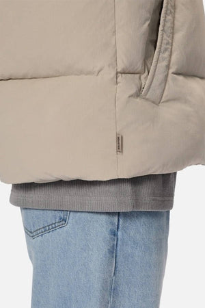 A close-up of a person wearing the Industrie The Busan Puffer Vest in beige nylon with Poly-fil padding over a gray sweatshirt and light blue jeans. Their hand is in the vest pocket against a plain, light background.