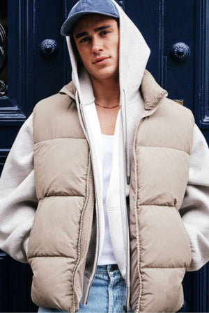 A young man stands before a dark blue door, wearing Industrie The Busan Puffer Vest with Poly-fil padding over a grey hoodie, white tee, blue jeans, and a blue baseball cap, looking confidently at the camera.
