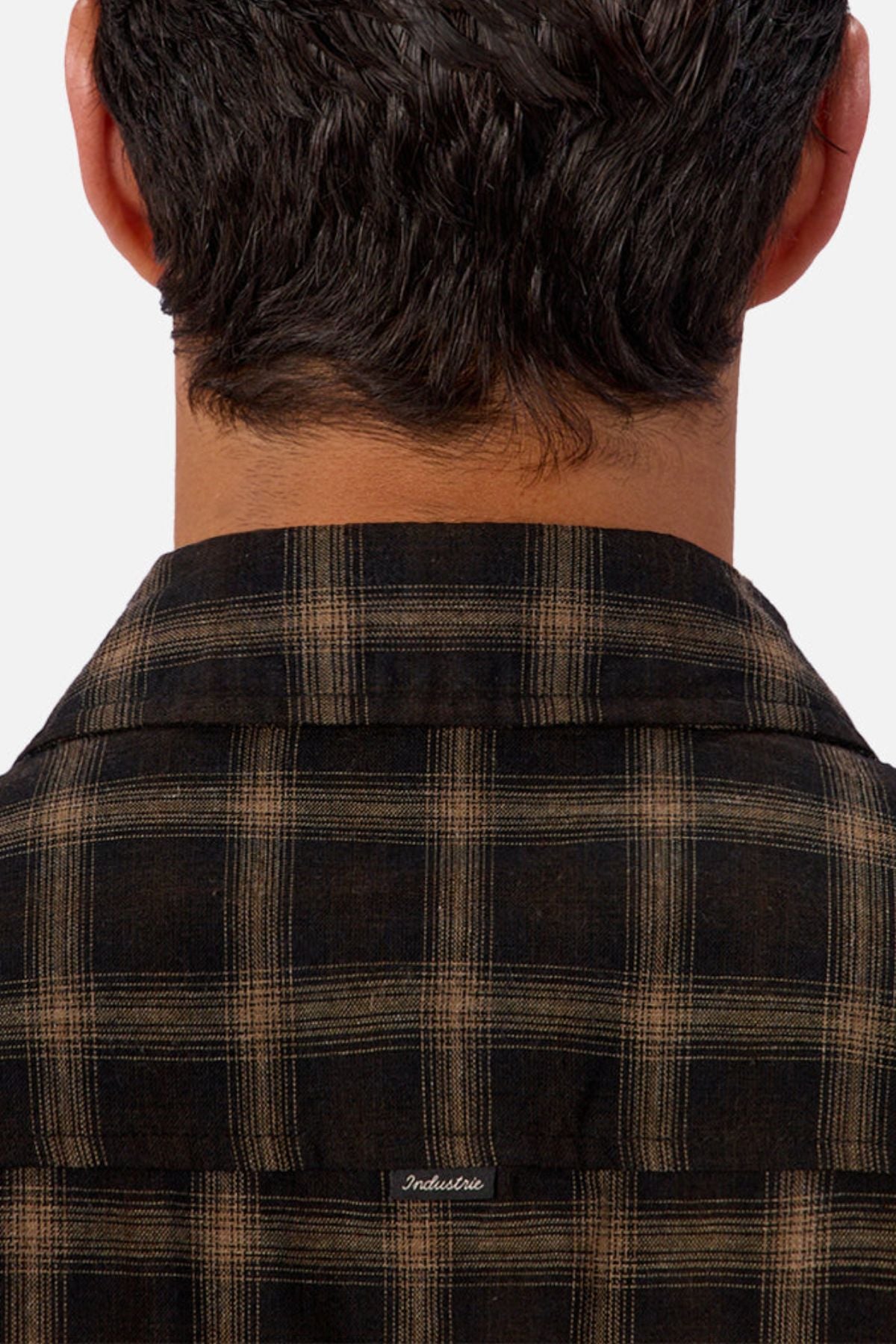 Close-up of the back of a person’s head, showing dark wavy hair and the collar of the Industrie The Carrington Shirt—a brown-and-black checked premium wool blend with "Indestructible" embroidered below the collar.