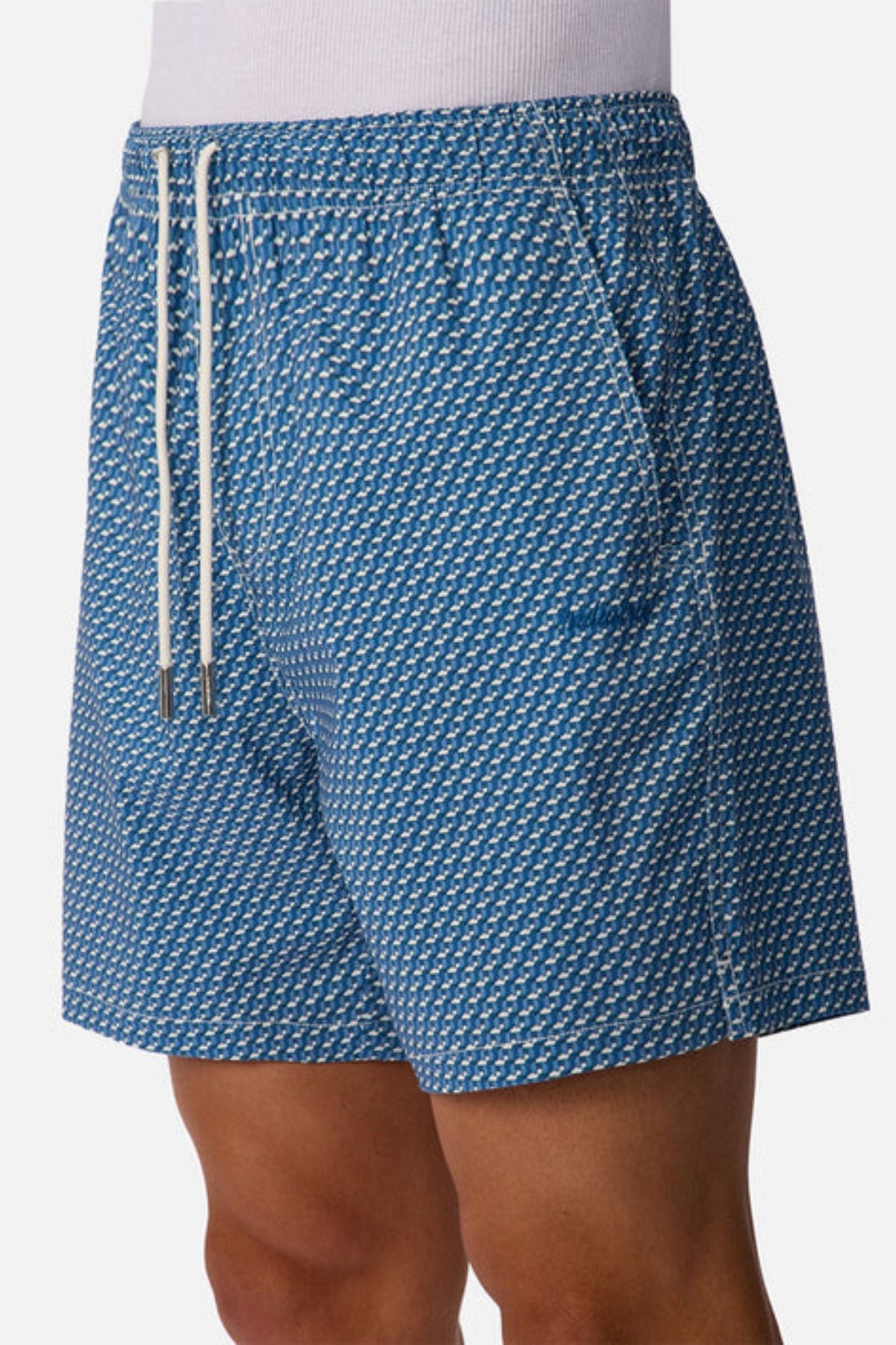 A person wears Industrie The Cayman Swim Short, featuring a subtle geometric abstract pattern, white drawstrings, and practical side pockets, standing against a plain white background.