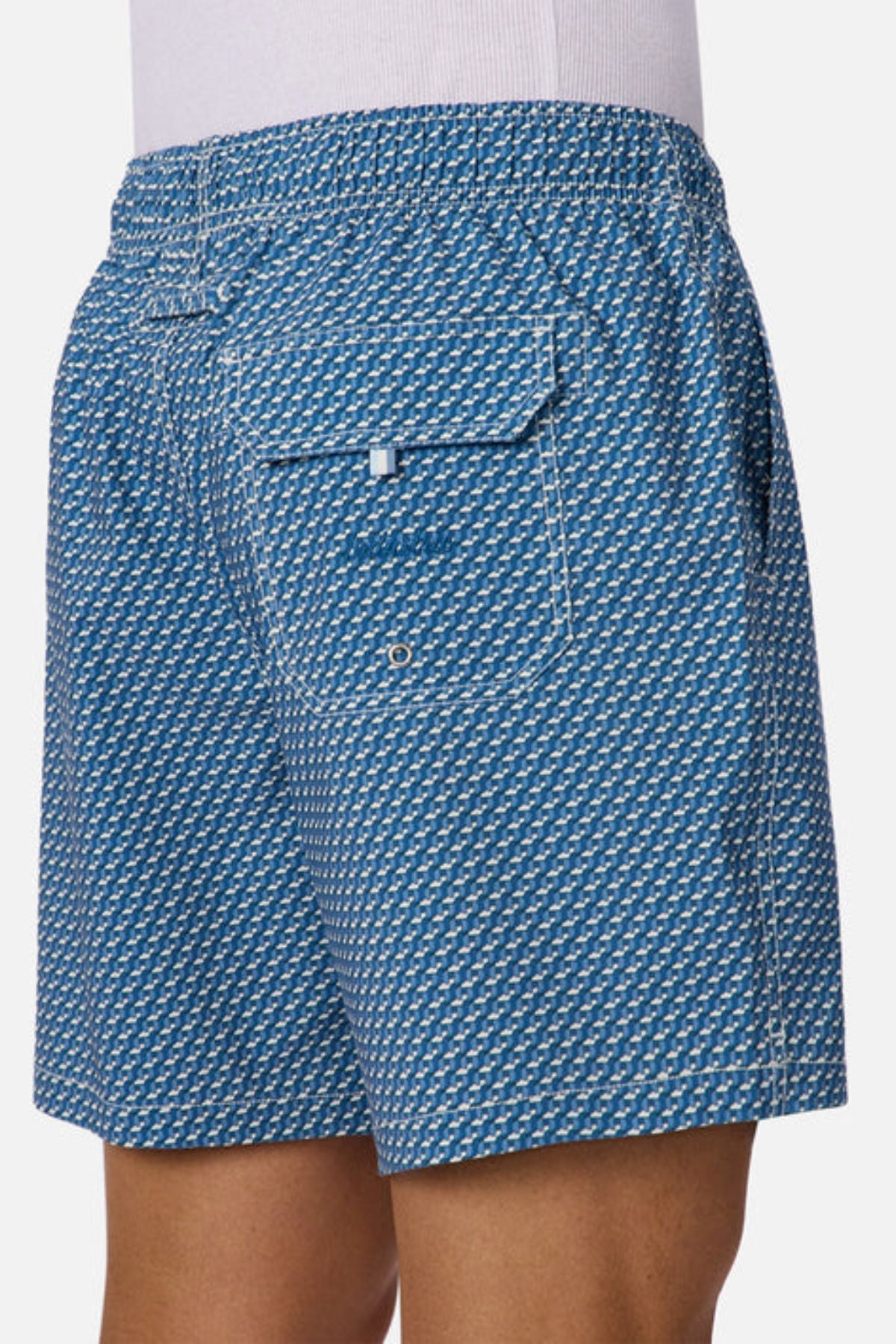 A person wearing Industrie The Cayman Swim Short, featuring a blue and white abstract pattern, back pocket, and white stitching, shown from the waist down and viewed from behind.