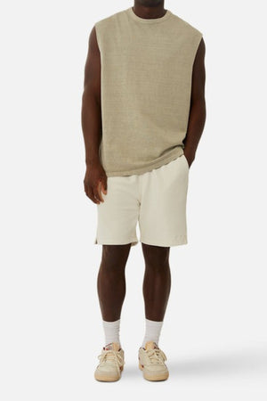 A person wearing the "Industrie The Del Sur Sleeveless Tee" in an oversized beige fit pairs it with white shorts, standing against a plain background. They complete the look with white socks and cream sneakers. This casual attire, crafted from 100% cotton, exudes a simple and relaxed vibe.