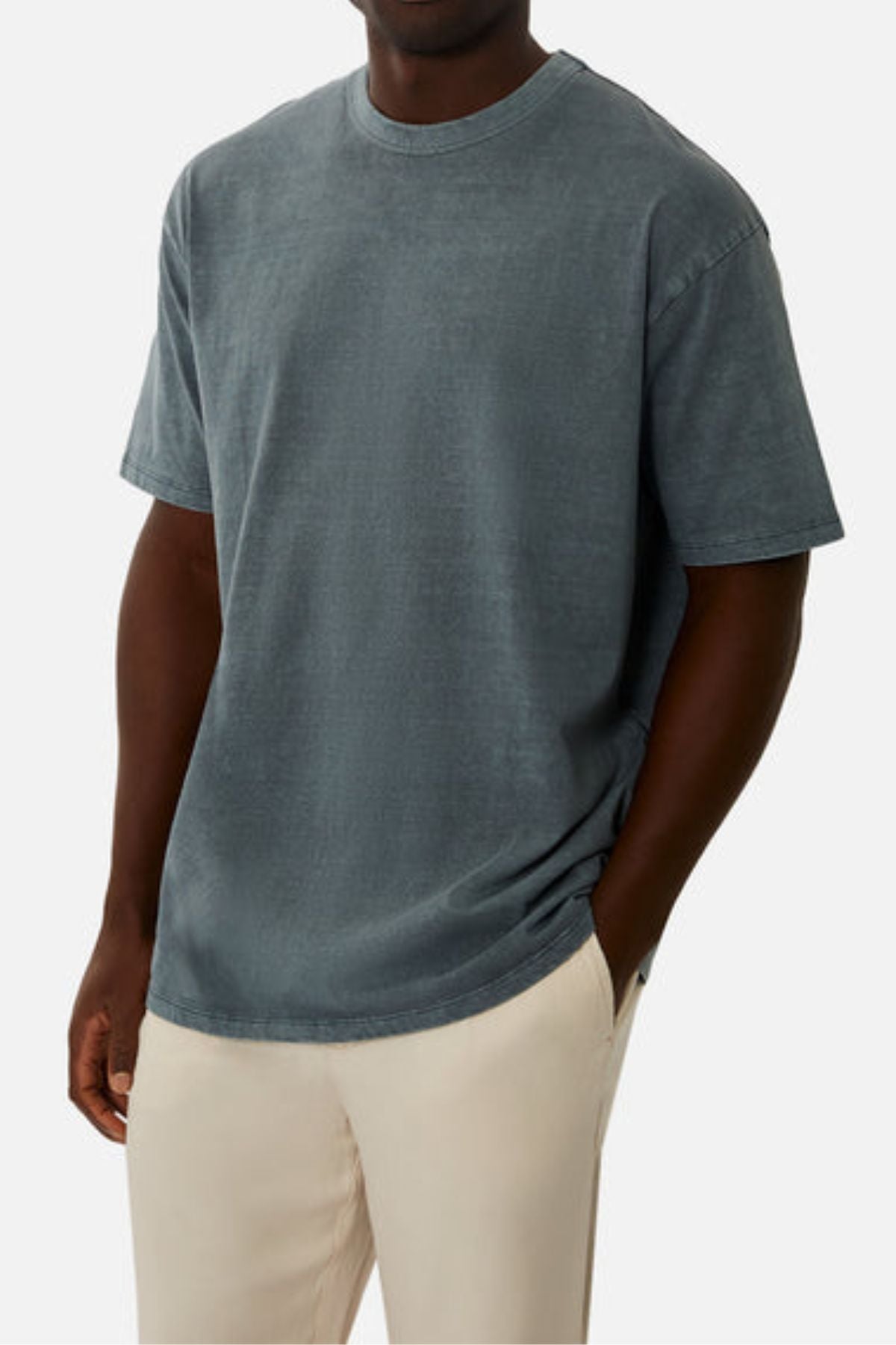 A man in an oversized, faded blue Industrie The Del Sur Tee crafted from heavyweight fabric and light beige pants stands with one hand in his pocket against a plain white background.