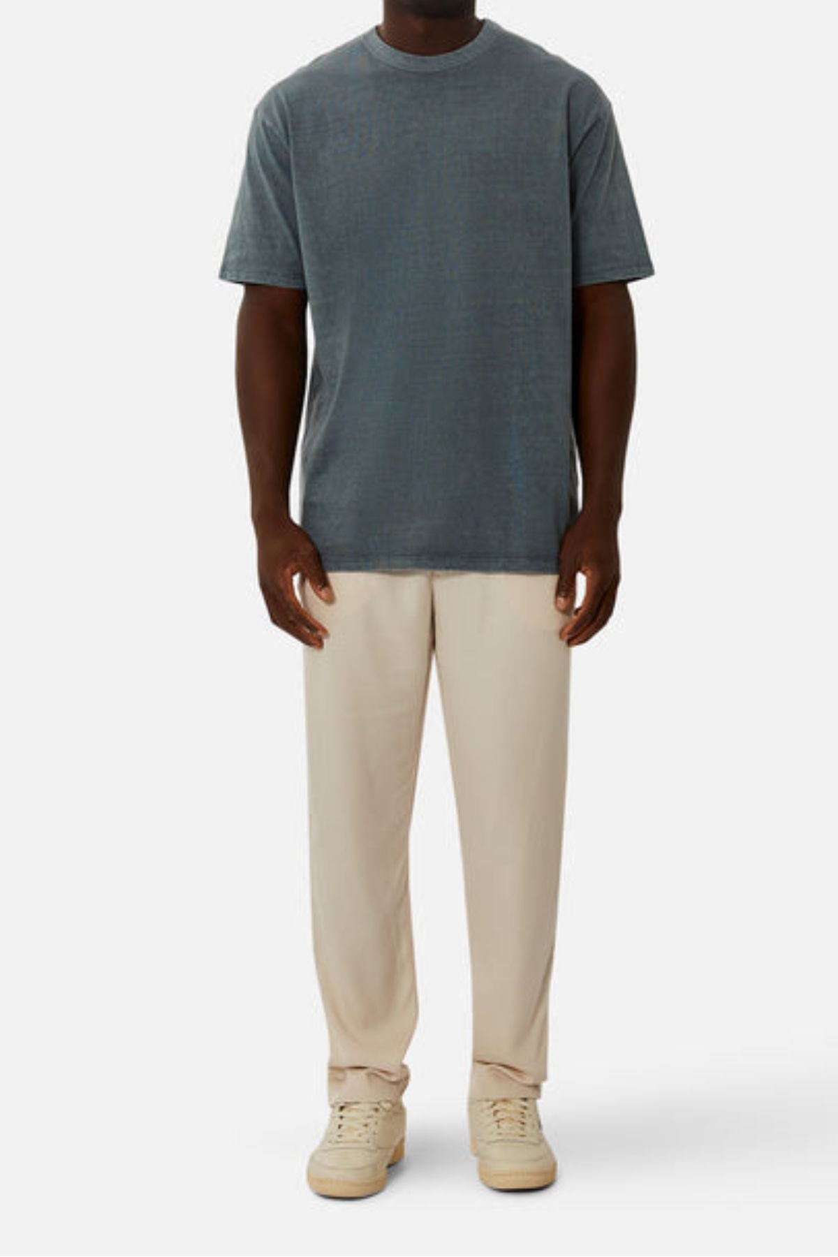 A person wearing an oversized fit faded blue Industrie The Del Sur Tee, light beige pants, and cream-colored sneakers stands facing forward against a plain white background.