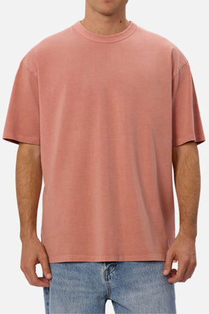 A person stands against a plain light background, dressed in the Industrie The Del Sur Tee. This oversized T-shirt features a faded pink hue and is made from heavyweight fabric with short sleeves, effortlessly pairing with light blue jeans for a relaxed-fit style.
