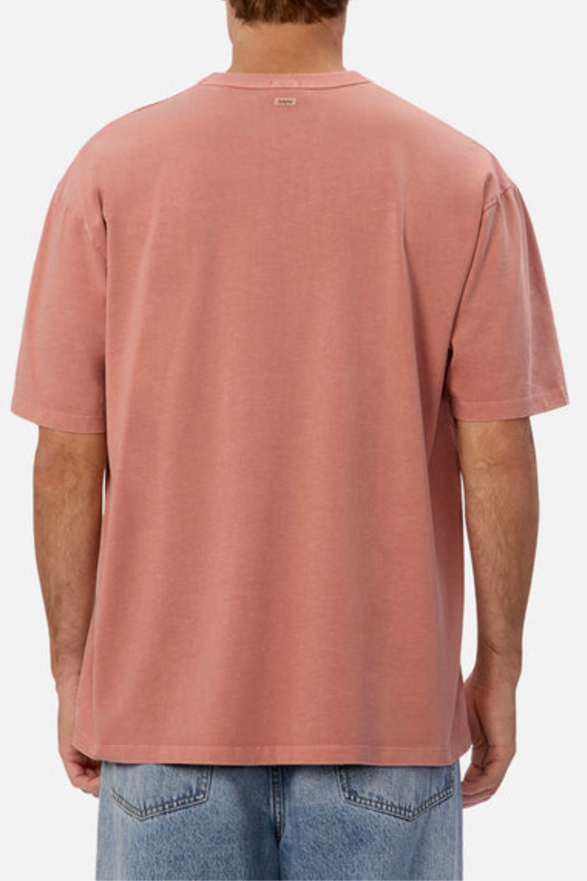 The image features an individual dressed in Industrie’s The Del Sur Tee in a pink color with an oversized fit, paired with light blue jeans. Captured from behind, the T-shirt showcases its short sleeves and heavyweight fabric, creating a comfortable and stylish appearance against a simple, light backdrop.