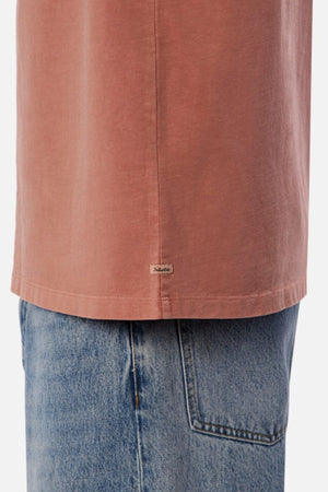 A close-up shows someone wearing the Industrie The Del Sur Tee in a faded pink shade, characterized by its oversized fit and a small tag near the hem. Teamed with light blue jeans, the heavyweight fabric and relaxed design offer an effortlessly casual appearance, drawing attention to the lower half of the tee and jeans.