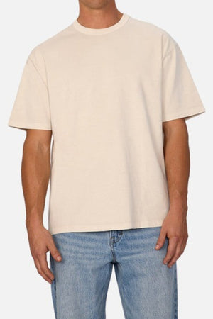 A person in the Industrie The Del Sur Tee—a plain beige short-sleeved T-shirt made from heavyweight fabric—and light blue jeans stands against a light gray background. Only their torso is fully visible, emphasizing the casual elegance of this laid-back ensemble.