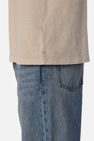 A close-up view features someone wearing the Industrie The Del Sur Tee, an oversized fit beige T-shirt made from heavyweight fabric, paired with blue jeans. The side of the T-shirt is visible alongside the hip area and pocket of the jeans, set against a neutral background.