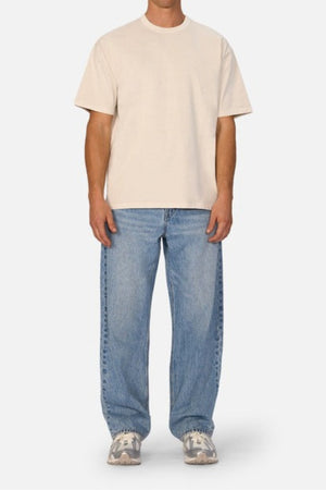 A person wearing an Industrie The Del Sur Tee, made from heavyweight fabric in a plain beige color, pairs it with loose blue jeans and white sneakers while standing against a neutral background. The outfit showcases an oversized fit for a casual and relaxed look.