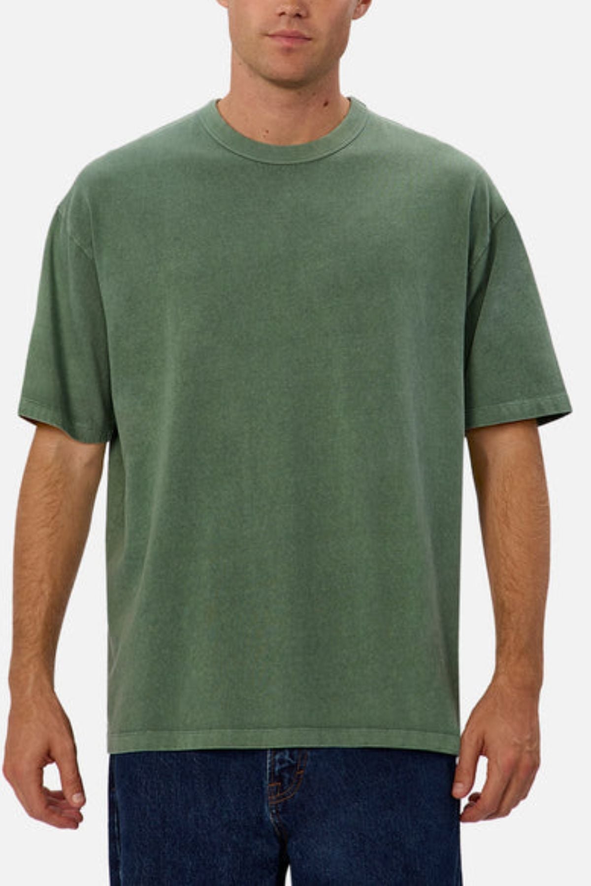 A person models the Industrie The Del Sur Tee, characterized by its oversized fit and heavyweight green fabric, set against a plain white backdrop. The tee features a straightforward round neckline and short sleeves, complemented by blue jeans.