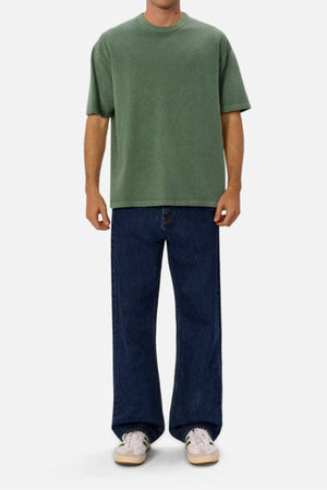 Wearing an oversized fit of the Industrie The Del Sur Tee in green, paired with dark blue wide-leg jeans and white sneakers, a person stands against a plain white background. The emphasis is on the relaxed, stylish look highlighted by the shirt's heavyweight fabric.