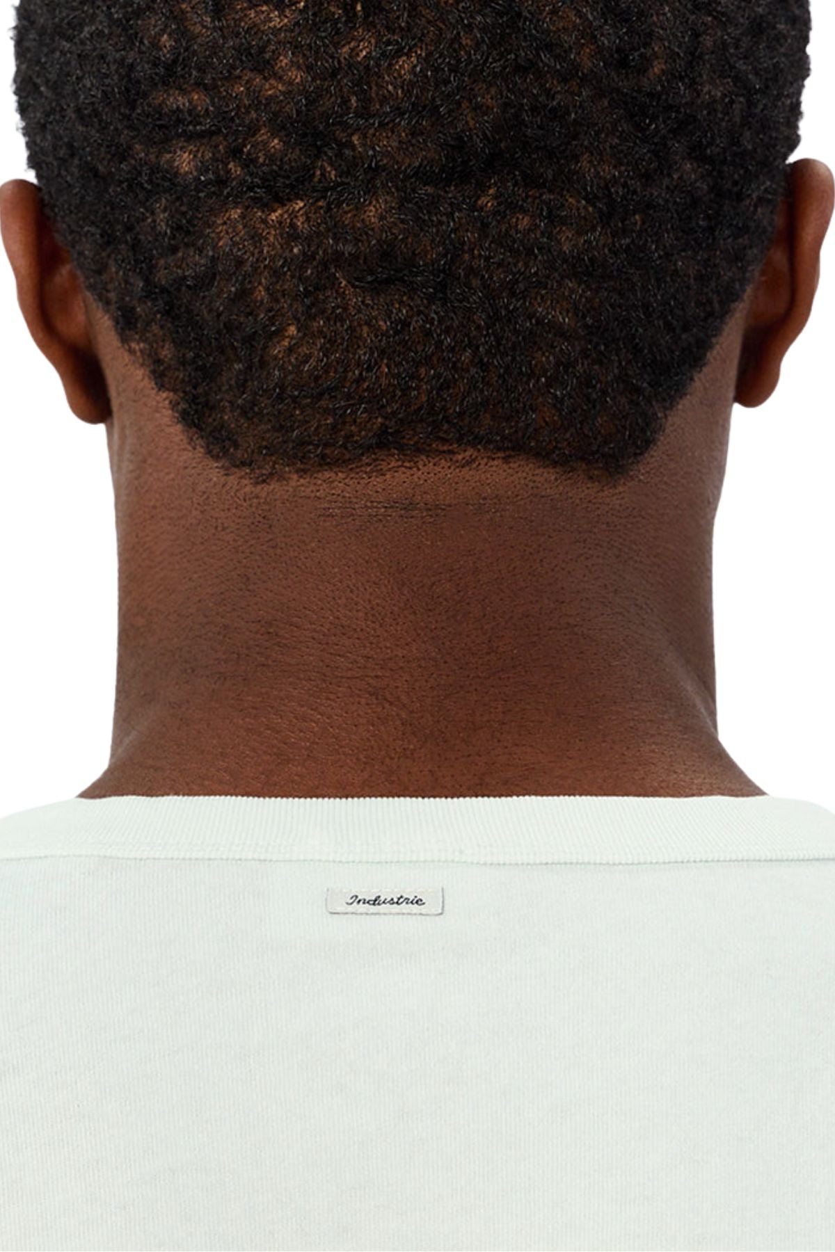 A close-up of the back of a person's head and neck reveals short, curly hair and the upper part of the light-colored Industrie The Del Sur Tee with a small "Industrie" label, showcasing its oversized fit.