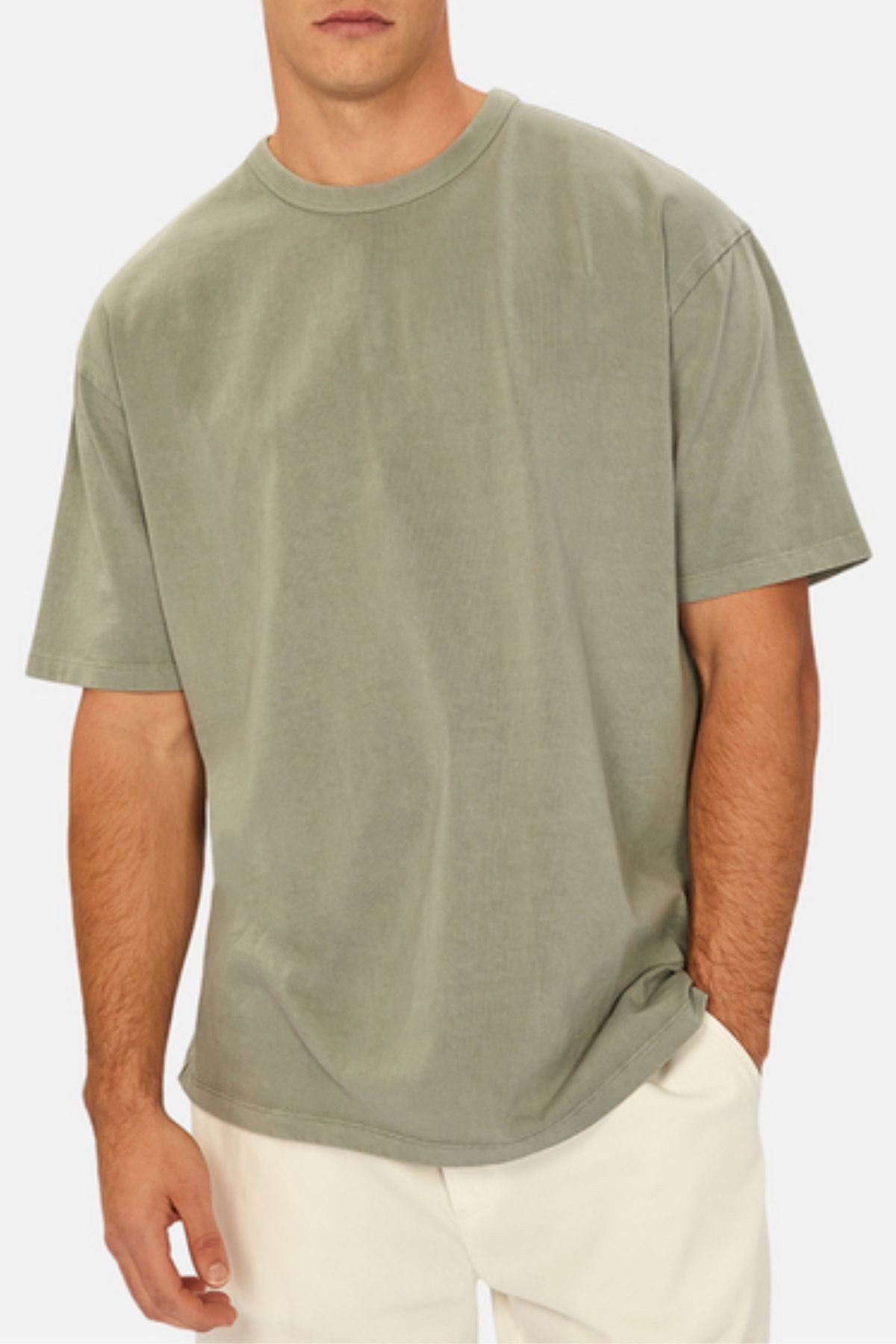 A man stands against a plain background wearing the Industrie The Del Sur Tee in light olive green heavyweight fabric and off-white pants, with one hand in his pocket.