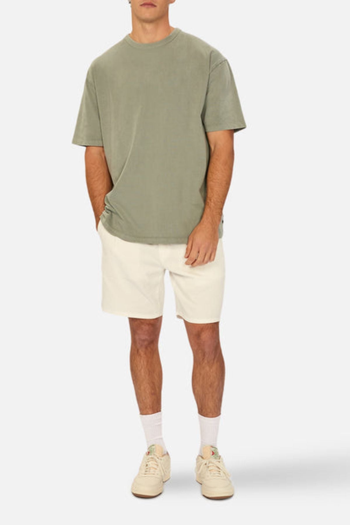 A man stands against a plain background wearing the Industrie The Del Sur Tee in olive green, paired with off-white shorts, white crew socks, and beige sneakers. His hands are in his pockets, and his head is cropped out of the image.