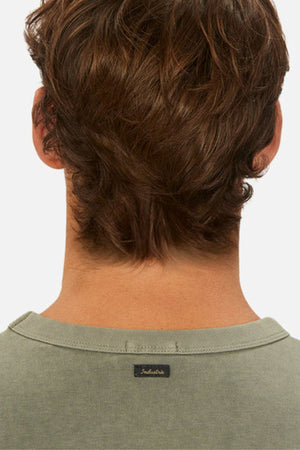 A person with brown, wavy hair is shown from behind in an oversized green Industrie The Del Sur Tee, made of heavyweight fabric. The shirt has a small black Industrie logo on the collar and is set against a plain white background.
