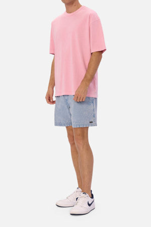 Wearing the Industrie The Del Sur Tee in pink, paired with light blue denim shorts and white sneakers featuring black accents, a person stands against a plain white background. The outfit showcases an oversized fit that exudes a casual and relaxed vibe.