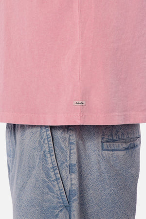 Close-up of a person wearing the Industrie The Del Sur Tee, an oversized fit pink shirt with a crew neck, paired with light blue jeans. A small rectangular tag is visible on the hem of the shirt. The heavyweight fabric of the jeans showcases a textured pattern and subtle pocket details.