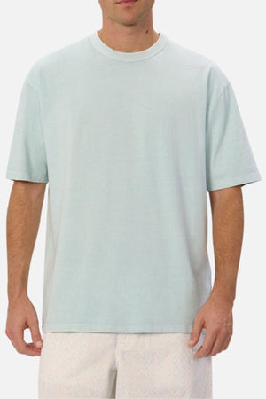 A man is dressed in an Industrie The Del Sur Tee in light blue, featuring an oversized fit, paired with light-colored shorts adorned with a subtle pattern. The tee's heavyweight fabric contrasts against the plain white background as he stands with his arms by his sides.